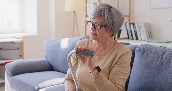 Lonely senior woman in eyewear having thoughtful sad expression, posing on sofa with walking stick, thinking about life, having health problems, needs support. Old people lifestyle concept