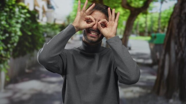 Man wearing gray turtleneck makes hand binocular gesture to eyes in a green park; exploration imagination joy curiosity.