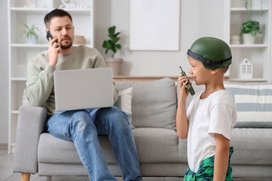 Little boy in soldier's helmet playing battle while his father talking by mobile phone and working with laptop at home