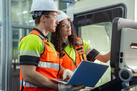 Two people wearing orange vests and hard hats are looking at a computer. Team of diverse engineers, male and female, programming and operating a CNC machine in a modern factory.