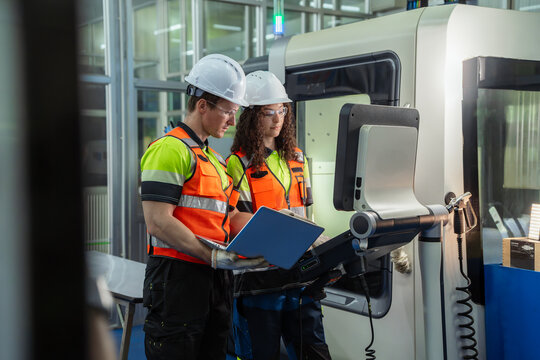 Two people wearing orange vests and hard hats are looking at a computer screen. Team of diverse engineers, male and female, programming and operating a CNC machine in a modern factory.