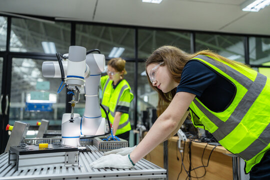 A woman in a yellow vest is working on a robot. Female students in a university robotics class learning to program a collaborative robot (cobot).