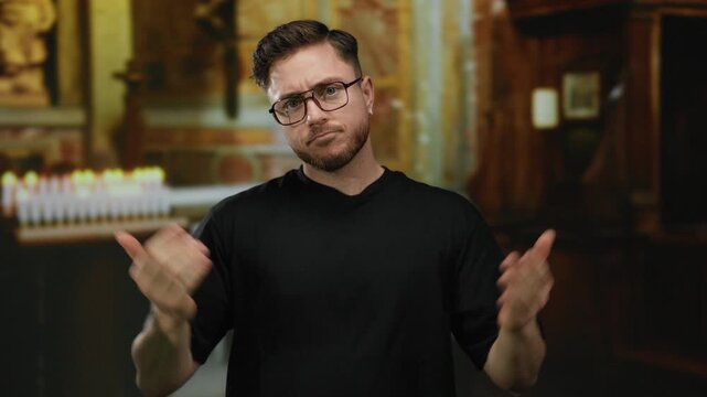 Young man with beard and glasses stands indoors in a church setting, appearing indecisive while looking at his palms in dimly lit surroundings with blurred candles in the background.