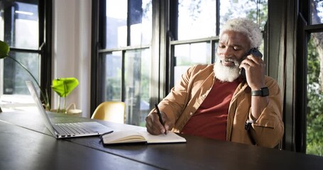 Senior African American man lifting phone, taking notes before typing on laptop at desk, copy space - Powered by Adobe