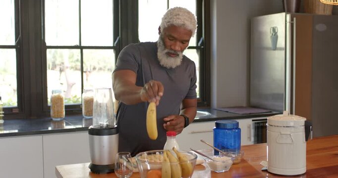 Senior African American man grabbing bottle then peeling banana at kitchen island for smoothie