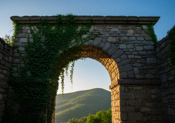 An old stone archway covered in ivy frames a scenic view of rolling mountains at sunset.