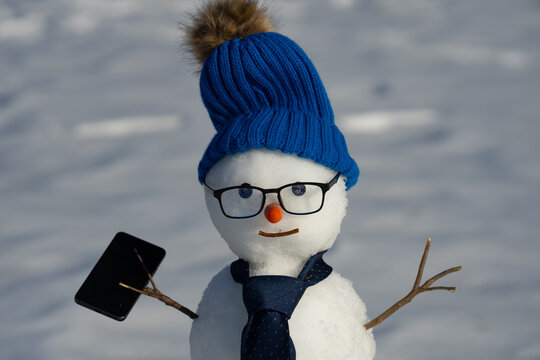 Business Snowman talking on smartphone in snowy landscape. Snowman making a business phone call. Funny snowman hacker using laptop outdoors. Snowman with mobile phone during winter celebration.