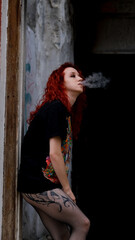 Fototapeta premium A young girl with curly red hair smokes a vape while in an abandoned old building. Young vaper. Bad habit. The difficult puberty period of a teenager.