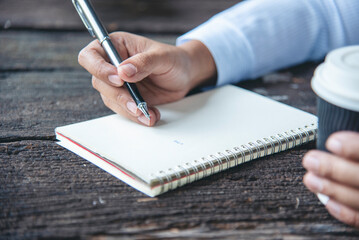 Woman hands writing empty notebook planing meeting agenda on wooden table. Close up women hands using pen drinking coffee cup planning office paper. Female hand holding pen note Personal Organizer