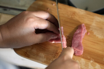 Close up of hands slicing raw meat into thin slices on a wooden cutting board with a sharp knife.