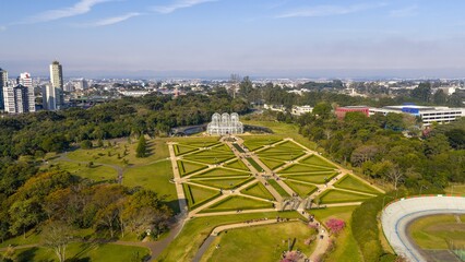 Oblique drone view of Curitiba Botanical Garden with symmetrical paths, greenhouse, and city in the background.