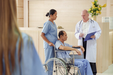 Fototapeta premium Senior asian male patient in wheelchair having medical consultation with senior caucasian male doctor and female nurse beside reception desk inside hospital lobby area