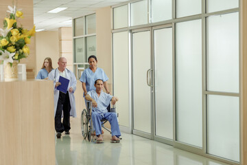 Medical team escorting asian male patient in wheelchair through hospital corridor including nurse, doctor and caregiver showing hospital teamwork during recovery and treatment support session