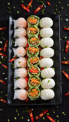 Colorful dim sum platter with shrimp dumplings, pork buns, and vegetable rolls, garnished with green onions and chili, isolated on black