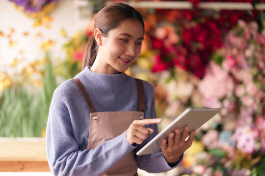 Asian adult woman florist busy managing flower shop orders using digital tablet for successful business growth delivering beautiful fresh arrangements daily - Powered by Adobe