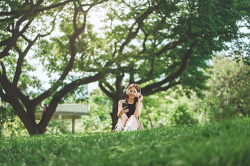 Cheerful asian woman wearing headphones and using smartphone to browse music or social media while sitting under a tree in city park during spring – relaxing lifestyle with soft sunlight, copy space