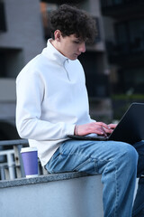 young male student with coffee to go sitting in park and using laptop while working on assignment