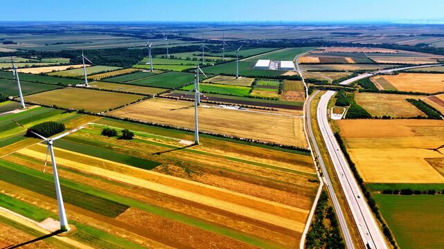 Green, yellow and brown patches of fields in the picturesque countryside. Wind turbines stand still on a windless day. Aerial view.