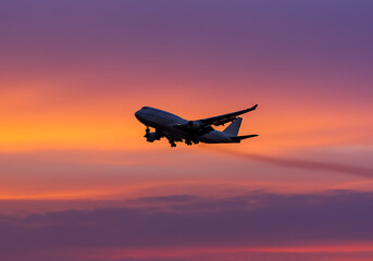 A large airplane is silhouetted against a vibrant orange and purple sky, ascending after takeoff at sunset.