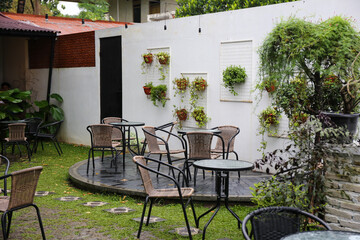A cozy outdoor seating area of a cafe with wicker chairs and tables. Lush green plants hang on a white wall, creating a natural and inviting atmosphere perfect for relaxation.