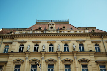 Facade of a historic yellow building with sculptures on the roof in the center of Zrenjanin, Serbia.