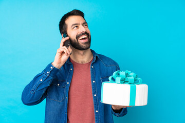 Young handsome man with a big cake over isolated blue background keeping a conversation with the mobile phone