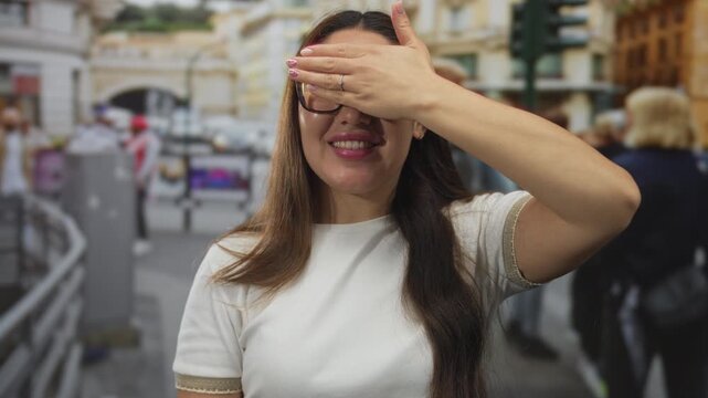 Young hispanic woman wearing glasses covers eyes with hand in front of building on busy urban street; curiosity.