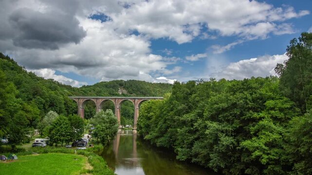 Historic railway viaduct spanning a verdant valley and reflective river in Herbeumont, Belgium. Top site surrounded by forest and peaceful campsite under  vibrant summer sky.