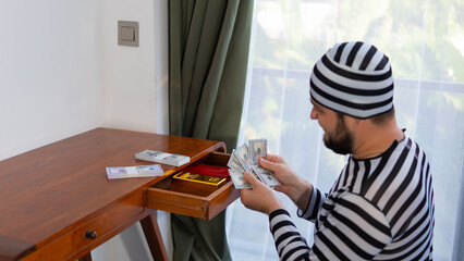 A man dressed as a cartoon burglar counts fake money beside a desk drawer filled with cash stacks...