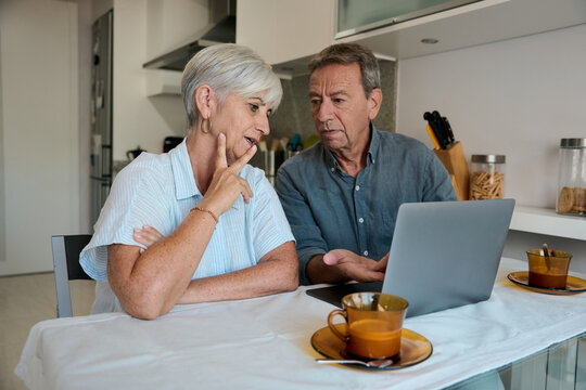 Senior couple using laptop at home having breakfast and discussing about finances
