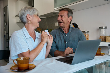 Senior couple making online purchases with credit card and laptop in kitchen