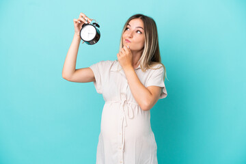 Young Romanian woman isolated on blue background pregnant and holding clock while thinking