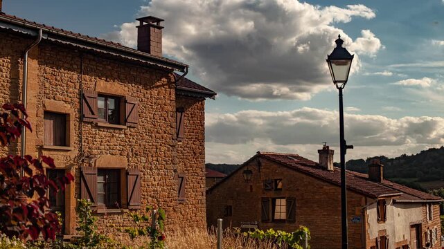 Warm sunlight bathes rustic stone houses with wooden shutters in Torgny, Belgium. A vintage lamppost and dramatic clouds add charm to this peaceful countryside village scene.
