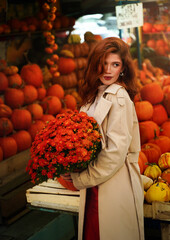 Beautiful red-haired young woman with orange bouquet of autumn flowers in her hands walks through the city vegetable market. Autumn and Halloween season.