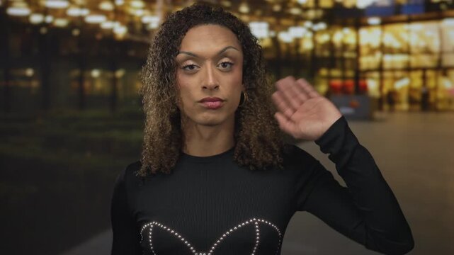 Transgender woman in black studded top waves hand on street under glowing streetlights at dusk; pride confidence visibility.