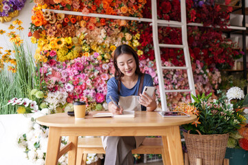 Asian female florist managing artificial flower shop business operations using smartphone and notebook for orders customer service and digital marketing