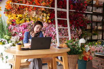Asian adult woman manages customer orders using laptop phone vibrant artificial flower boutique interior business owner entrepreneur shop worker