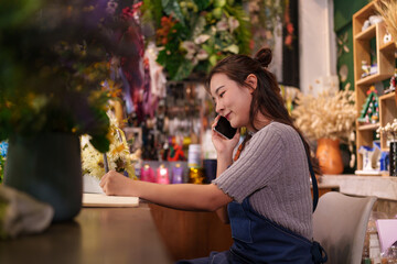 Young Asian woman flower shop owner manages business calls writing notes for customer orders in beautiful artificial floral store