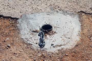A steel anchor tube set in concrete with an attached metal chain is embedded in the ground, used to securely fasten or lock equipment, barriers, or structures outdoors