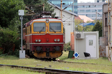 Fototapeta premium A vintage red and yellow electric locomotive stands on a railway siding surrounded by greenery and utility buildings, reflecting classic European rail transport design
