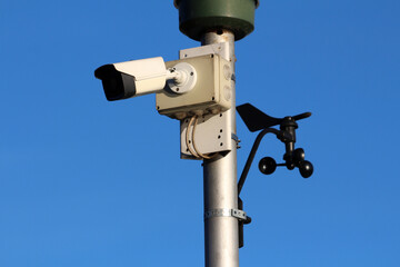 A metal pole supports a white surveillance camera and black anemometer, capturing traffic and weather data, ideal for monitoring road safety in urban or rural environments
