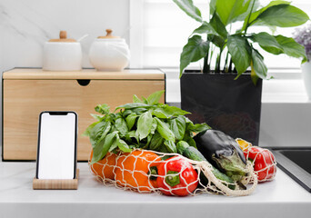 Hand with white screen phone near fresh vegetables and fruits in a reusable bag in the modern kitchen. Food delivery. Grocery shopping online. Eco-Friendly Shopping. The idea of a healthy lifestyle