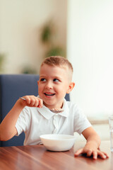 Cute 5-year-old boy having breakfast at home, eating porridge with water. Natural light, healthy food