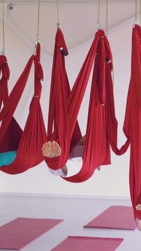 sleepy women group with bare feet relaxes swinging in red aerial fly yoga hammocks above fitness sport mats