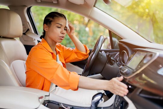 Frustrated young black woman inside car feeling hot, trying to adjust settings on dashboard. African american female checking temperature of air condition in vehicle