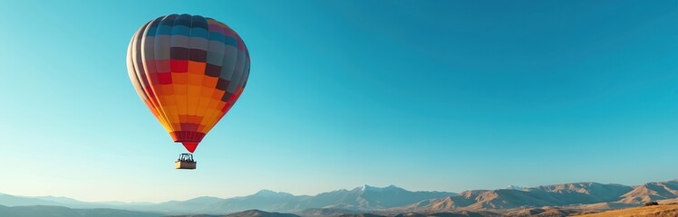 Naklejka premium Colorful hot air balloon glides across vibrant blue sky over mountainous landscape. Experience freedom, adventure, exploration. Enjoy peaceful soaring journey, breathtaking views, majestic voyage