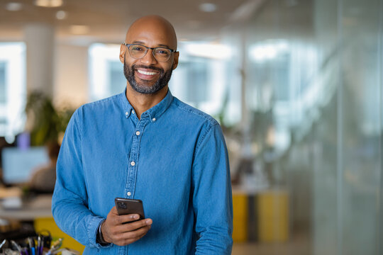 Smiling man in glasses and denim shirt holding a smartphone in a modern office, standing confidently