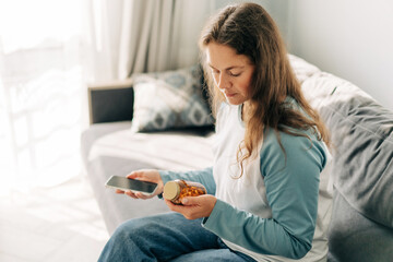 Woman holding pills and mobile phone while sitting on sofa in living room.