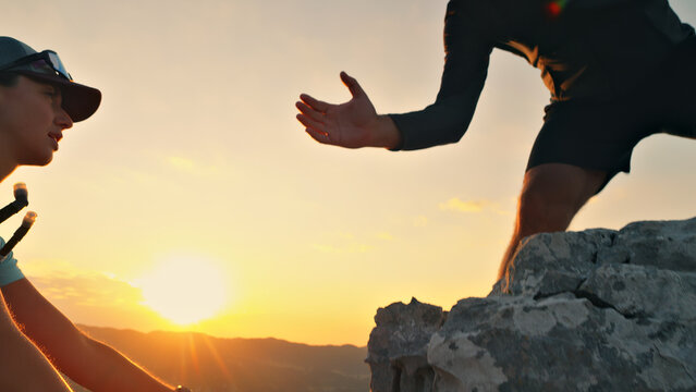 Two runners at sunset on a rocky peak, one helping the other, symbolizing teamwork and endurance in cross-country running. Golden light highlights their mutual support and achievement.
