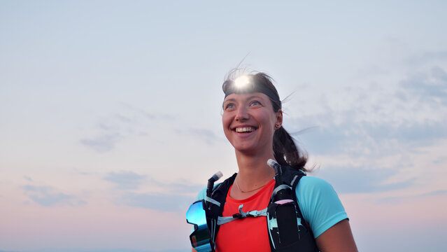 Joyful Caucasian woman in athletic gear, headlamp, and hydration pack, smiles triumphantly against a twilight sky, embodying cross-country running and endurance.